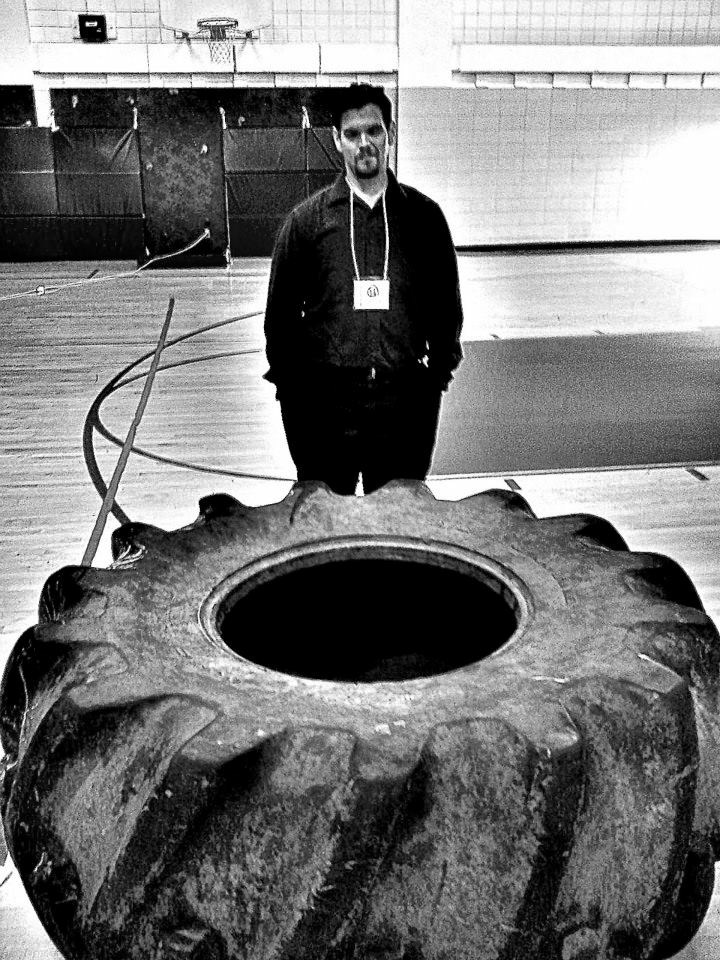Justin standing behind a large tire in a school gym